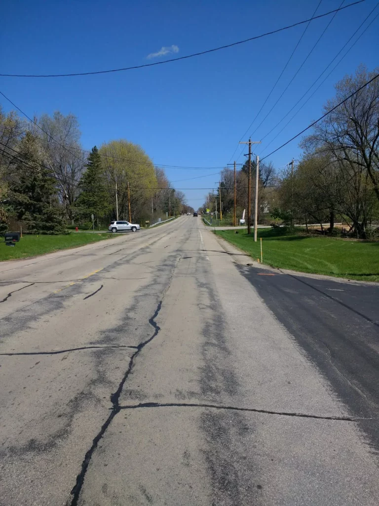 Long residential road with cracks surrounded by grass, trees, electrical poles and a car in the distance
