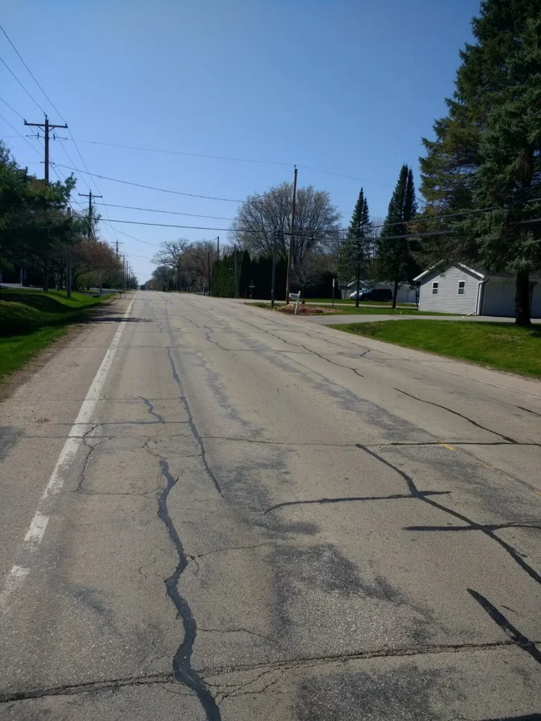 Road with cracks surrounded by grass, trees and a house