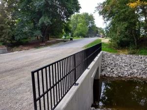 Water runs under a small bridge with a metal railing in an area surrounded by trees
